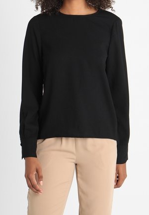 Kobieta ubrana w gładką czarną bluzkę na długi rękaw i beżowe spodnie, stojąca z ramionami swobodnie opuszczonymi wzdłuż ciała na białym tle.