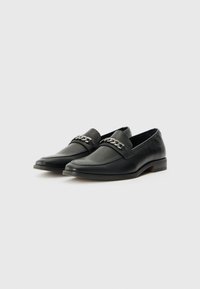 Mocassins noirs en cuir lisse, dotés d'un accent de chaîne en argent décoratif sur le devant, avec un talon bas empilé et des détails cousus.