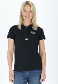 Sort polo shirt med krave, med hvid kant, knapper og et logo på venstre bryst. Båret med blå jeans.