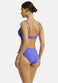 Vrouw met een knot in een paarse bikini, gezien van achteren, waarbij het ontwerp en de pasvorm van de zwemkleding tegen een effen achtergrond worden getoond.