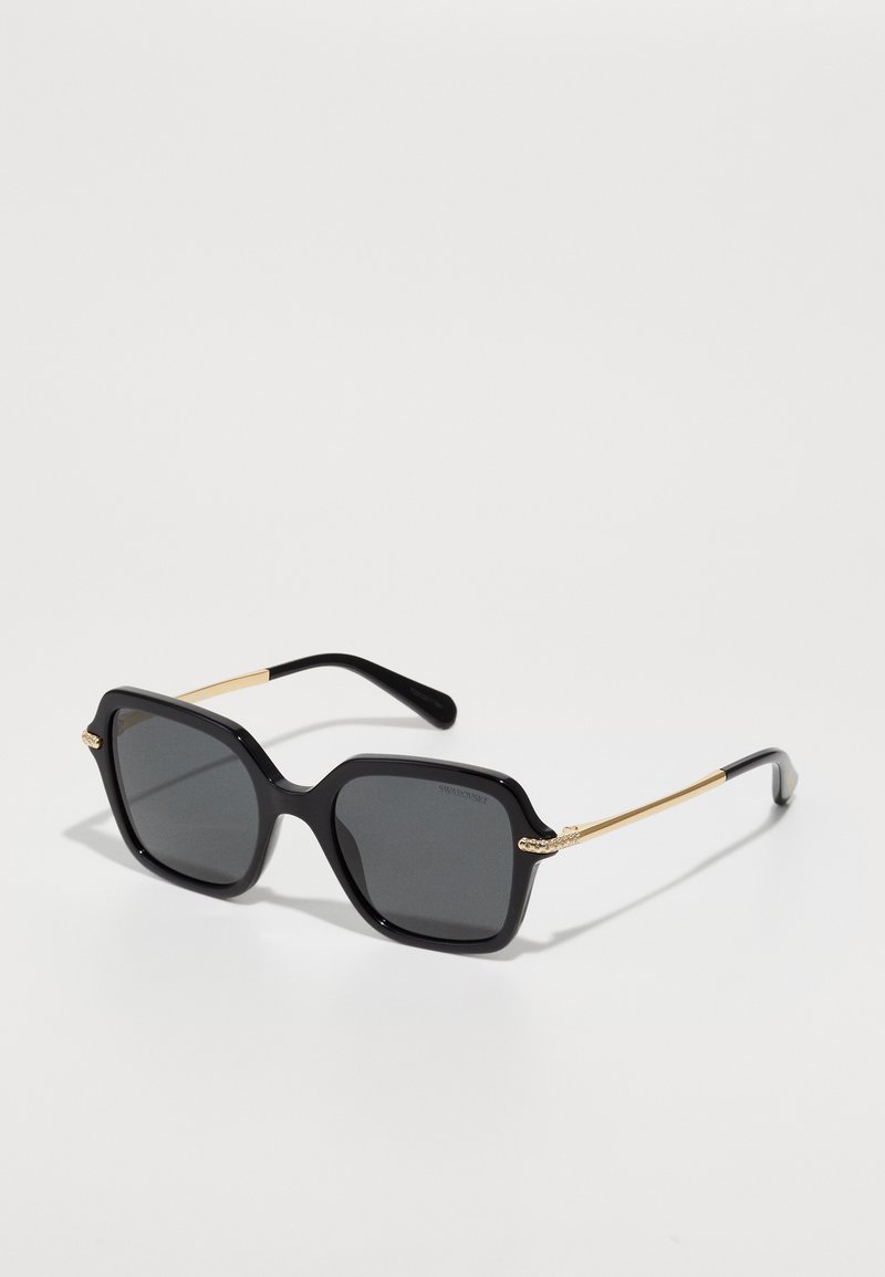 Occhiali da sole oversize neri con montatura spessa, lenti scure e dettagli dorati sulle tempie. Presentano una forma geometrica e una texture liscia.