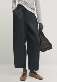 Pantalons larges marine à plis, associés à un haut ajusté à rayures à manches longues ; les accessoires comprennent un sac en daim marron foncé et des chaussures marron clair.