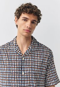Camicia a maniche corte in tessuto leggero con motivo a quadri con quadrati blu scuro, arancioni e bianchi. Colletto classico e chiusura con bottoni.