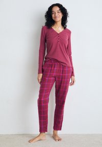 Mørkerød langermet topp med henley-hals. Rutete pysjshorts i mørkerød og rosa med elastisk liv. Modell står barbeint på teppet.