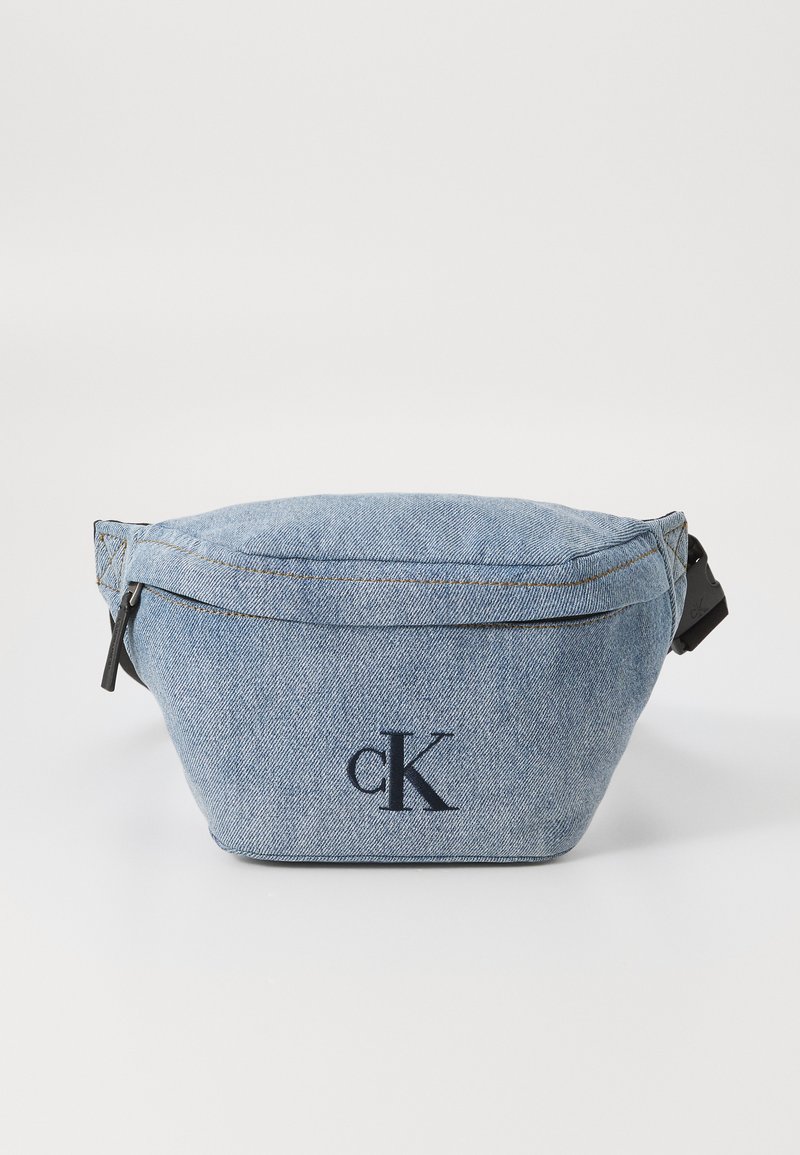 Denim fanny pack with a rounded shape, blue color, visible stitching, and a black zipper. Features a prominent embroidered logo on the front.