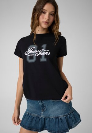 Ung kvinde iført en sort Guess Jeans t-shirt med "81" print og et blåt denim-flæsekørt mod en ensartet lys grå baggrund.