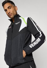 Black and white athletic jacket with green accents, high collar, padded chest area, and prominent "VENUM" branding on sleeves. Zipper closure.