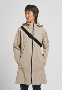 Beige wasserfester Mantel mit Kapuze, langen Ärmeln, Frontreißverschluss und seitlichen Taschen; verfügt über einen schwarzen verstellbaren Gurt quer über den Körper.