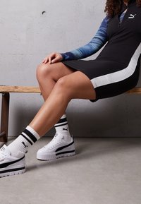 Abito nero con strisce bianche sui lati, maglietta a maniche lunghe blu, sneakers bianche con dettagli neri e calze a righe. Sfondo con panchina in legno.