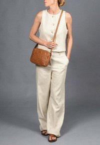 Beige linnen top zonder mouwen met drie knopen, gecombineerd met losse beige broek en een bruine gevlochten schoudertas. Bruine sandalen maken de look af.