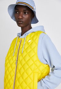 Veste jaune matelassée avec une fermeture éclair argentée, portée par-dessus un hoodie bleu clair. Un chapeau bucket bleu clair présente un accent rayé et un détail de logo.