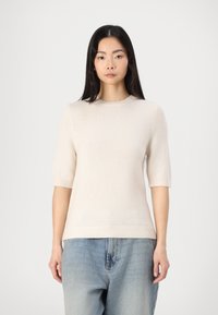 Strickpullover in hellbeige mit kurzen Ärmeln, rundem Halsausschnitt und geripptem Saum. Getragen mit lockeren blauen Jeans aus Denim, die eine verblichene Textur aufweisen.
