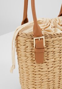 Borsa in paglia intrecciata con fodera in tessuto crema arricciato, manici in ecopelle marrone, dettaglio della fibbia dorata e design a apertura aperta.