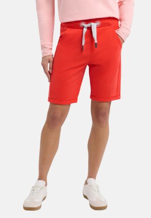 Person trägt leuchtend rote, knielange Shorts mit weißem Kordelzug, pinkes Langarmshirt, weiße Sneakers, Hände in den Taschen.