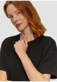 Juoda trumpomis rankovėmis palaidinė su tekstūrine šešiakampių raštu, plačia iškirpte ir švelnia medžiaga. Modelis su raudonais plaukais laiko ranką prie kaklo.