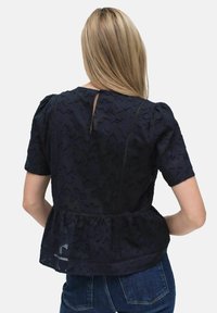 Sininen, teksturoitu paita lyhyillä puffihihnoilla, peplum vyötäröllä ja takana on avaus, jossa on avaimenreikä. Materiaali sisältää kohokuvioinnin.