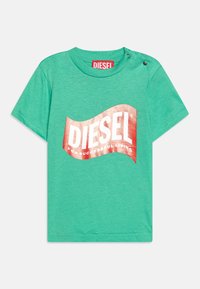 Zelené krátké tričko s krátkým rukávem vyrobené z bavlny. Na přední straně se nachází velké kovové červené logo "DIESEL" ve vlnovém tvaru. Na rameni je detail se zapínáním.