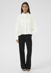 White, semi-sheer blouse with a collar, front button closure, and long sleeves, paired with black, loose-fitting trousers.