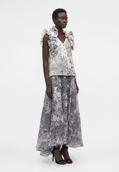 Γυναίκα φοράει μπλούζα χωρίς μανίκια με λουλουδένιο σχέδιο και βολάν στους ώμους, καθώς και μακριά διαφανή φούστα με λουλουδένιο μοτίβο, συνδυασμένη με μαύρα ψηλοτάκουνα σανδάλια.