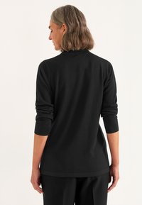 Frau mit schulterlangem Haar, die ein schwarzes Langarmoberteil und schwarze Hosen trägt, steht mit dem Rücken zu einem schlichten Hintergrund.