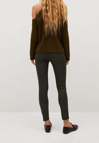 Mujer con largo cabello rubio ondulado que lleva un suéter verde oliva con hombros al descubierto, pantalones oscuros ajustados y zapatos negros tipo slip-on, de pie mirando hacia atrás.