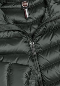 Veste matelassée vert foncé avec une fermeture éclair partiellement ouverte et une étiquette noire Colmar Originals avec une patte en tissu rouge, blanche et grise à l'intérieur du col.