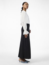 Blouse blanche avec des détails en dentelle sur les manches, associée à une longue jupe noire. Chaussures à talons bleues avec un bout pointu.