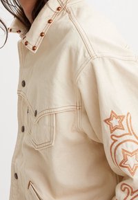 Béžová džínová bunda s měděnými cvoky na límci a oranžovou výšivkou hvězd na rukávu; obsahuje nasazovací kapsy a kontrastní stehy.