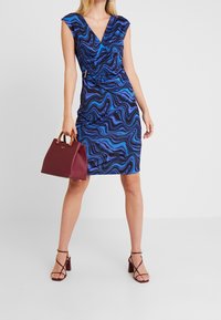 Blå och svart mönstrad klänning med V-ringning, rynkad design, bärs med en vinröd handväska och remsandaler. Slät, stretchig tyg.