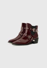 Bottines en cuir verni bordeaux avec des pointes triangulaires, deux sangles réglables, accessoires en argent et un petit talon carré.