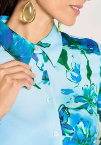 Woman wearing large gold teardrop earrings and a light blue button-up shirt with blue and green floral patterns.