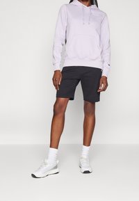 Person klädd i en ljuslila Champion-hoodie, svarta shorts, vita strumpor och vita sneakers som står mot en enkel bakgrund.