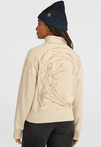 Beige sweatshirt med hög krage och en surfdesign i brunt på baksidan. I bomullstyg med ribbade muddarmar, tillsammans med en mörk mössa.