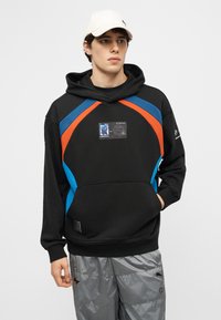 Jeune homme portant un sweat à capuche noir avec des rayures bleues et orange, un écusson graphique sur la poitrine, un pantalon argenté réfléchissant et une casquette blanche, debout immobile.