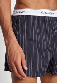 Tumši zili kokvilnas bokseršorti ar vertikālām baltām svītrām, ar baltu jostu, uz kuras redzams "Calvin Klein" logo, un priekšējo pogu aizdari.