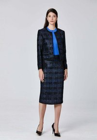 Costume à motifs bleu marine et noir avec une veste ajustée et une jupe crayon, associé à un chemisier bleu. Texture lisse avec des escarpins noirs élégants.