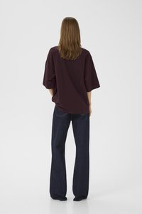 Person med langt brunt hår står med ryggen til, iført en løs mørke lilla t-skjorte med korte ermer og vide mørkeblå jeans.