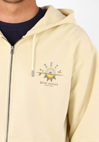 Sweat à zip couleur jaune clair en tissu doux, avec un logo sur le devant au thème soleil et surf, et capuche à cordon.