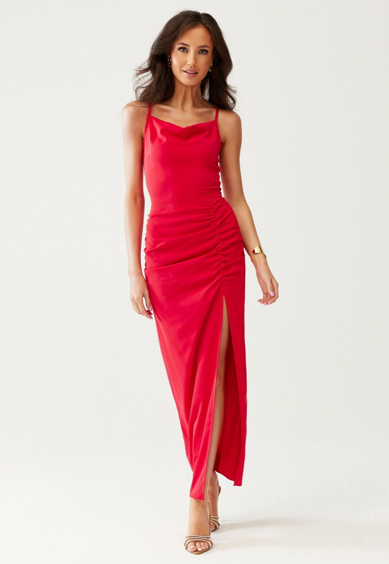 Vrouw in een rode mouwloze jurk met ruches en een zijsplit, met gouden oorbellen, een armband en beige hoge hakken, poseert tegen een witte achtergrond.