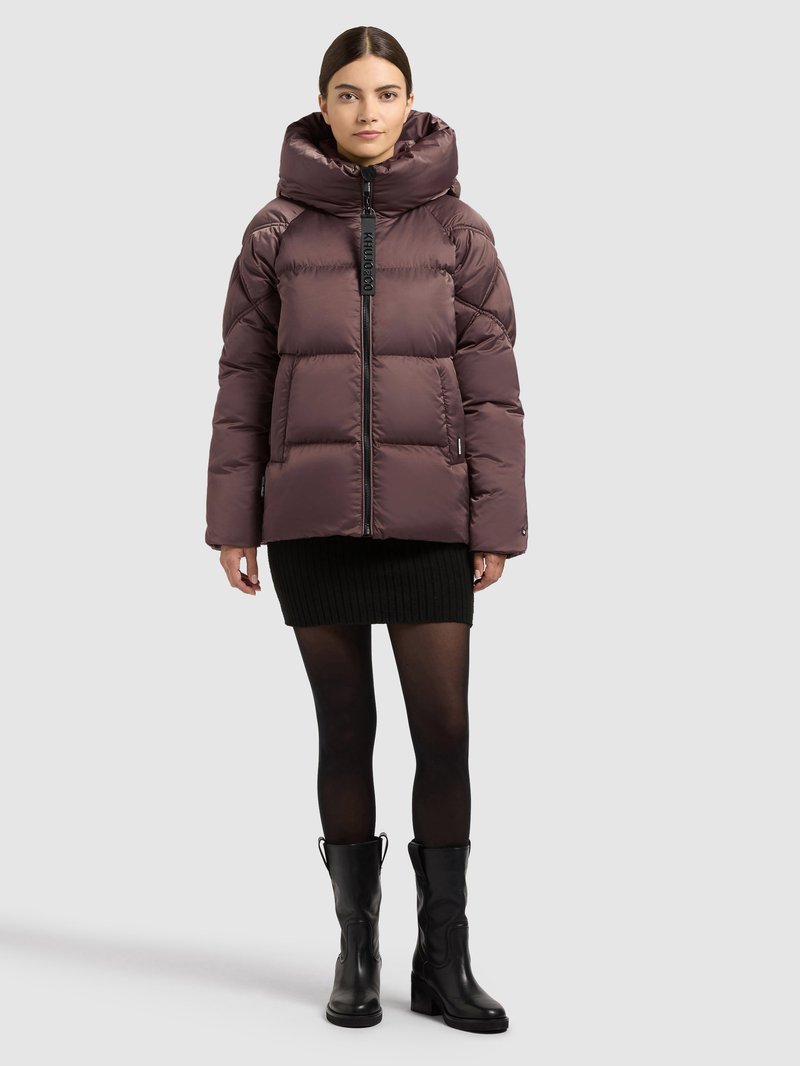 Polstret lilla jakke med høj krave, lynlås og sidelommer, båret over en sort ribbet nederdel og sorte støvler til knæet.