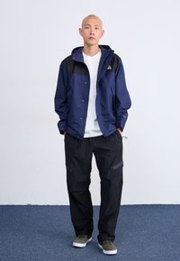 Tamsiai mėlyna vandeniui nepralaidi striukė su juodais akcentais, kapišonu su raišteliais ir spaustukų užsegimu; derinama su laisvo silueto juodomis kelnėmis ir žaliais sportbačiais.