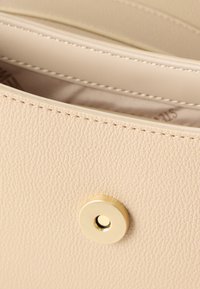 Μπεζ τσάντα χειρός από δερμα με υφή, με ορατή ραφή και στρογγυλό χρυσό μαγνητικό κούμπωμα που είναι μερικώς ανοιχτό, αποκαλύπτοντας τη φόδρα του εσωτερικού.