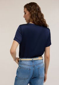 T-shirt in satin blu navy con maniche corte, caratterizzato da un collo rotondo e risvolti sulle maniche. Abbinato a jeans in denim blu chiaro, con tasche posteriori in evidenza.