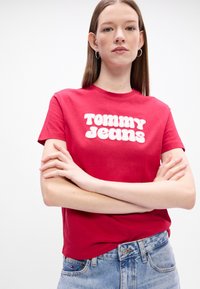 Punane puuvillane T-särkk, lühikeste varrukatega, millel on rinnal suur valge kiri "TOMMY JEANS". Mudel kannab kergeid siniseid teksaseid.