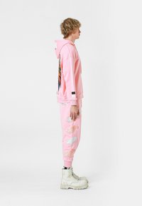 Sudadera rosa con un gran gráfico colorido en la parte posterior, combinada con pantalones de chándal rosa a juego con patrones de plumas bordados. Botas blancas de suela gruesa.