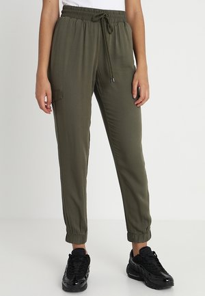 Oliwkowe spodnie dresowe z elastycznym pasem i mankietami przy kostkach, boczną kieszenią, noszone z czarnymi sneakersami i białą bluzką.