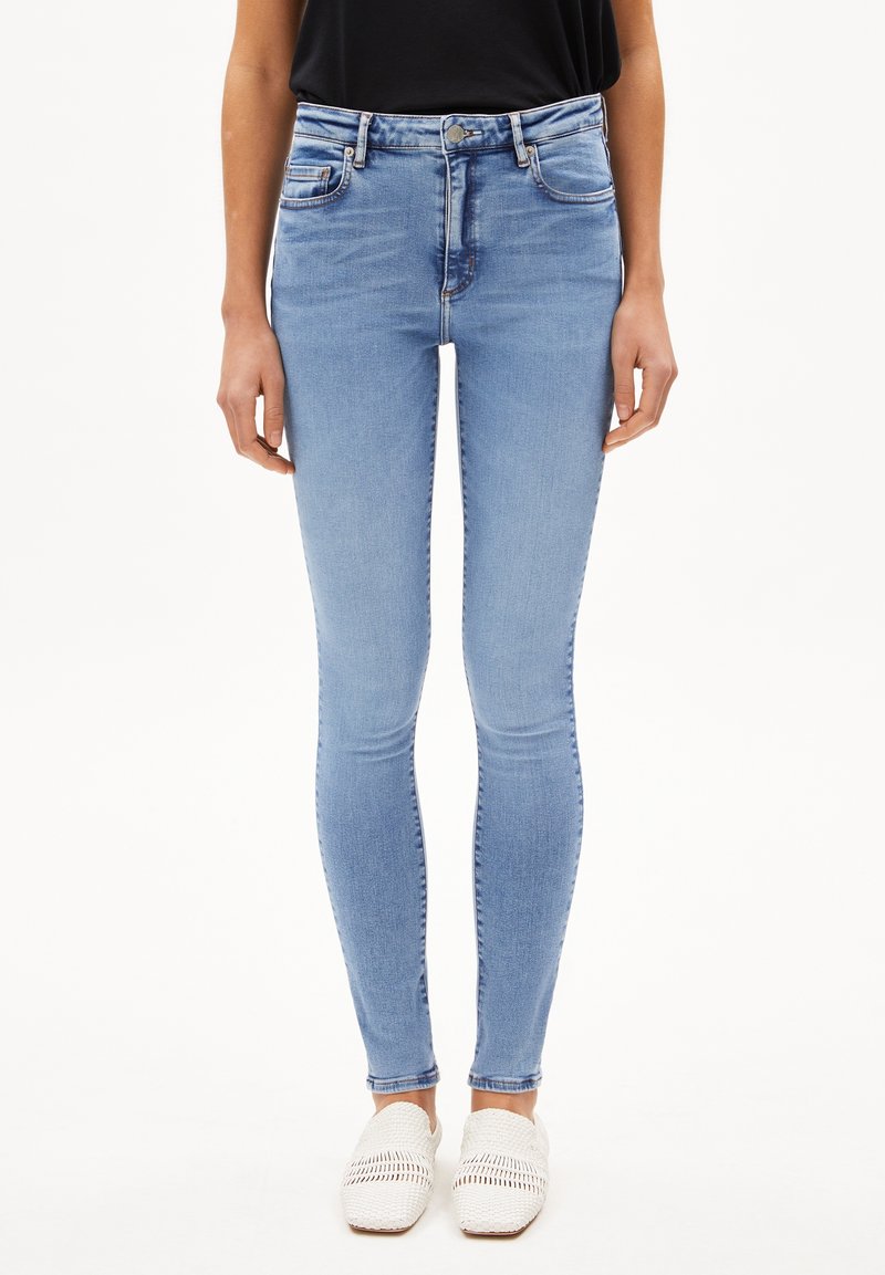Femme portant un jean skinny bleu clair à taille haute et des chaussures blanches tissées sans lacets, debout devant un fond uni.