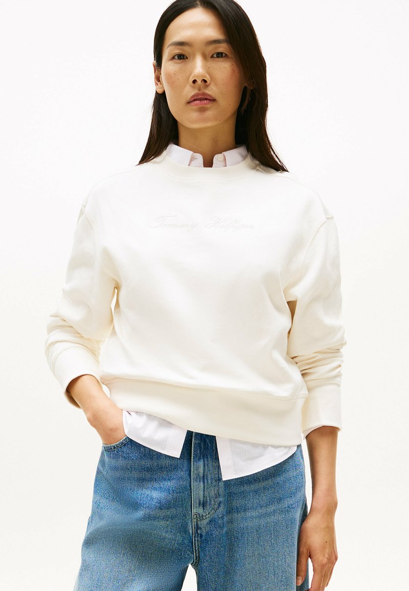 Sweatshirt color crema con collo tondo, polsini a costine e logo ricamato. Indossato sopra una camicia bianca con colletto e jeans blu.