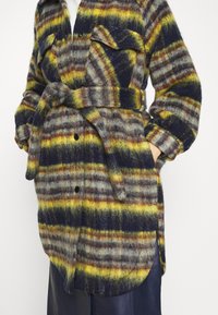 Long manteau duveteux à rayures horizontales jaunes, grises, brunes et noires, ceinturé à la taille, porté sur un pantalon foncé.
