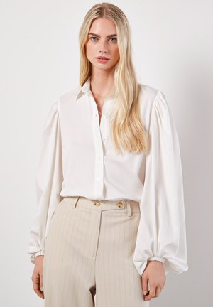 Blusa bianca a maniche lunghe in tessuto liscio, con chiusura frontale a bottoni, maniche a sbuffo e colletto classico. Abbinata a pantaloni beige a righe.